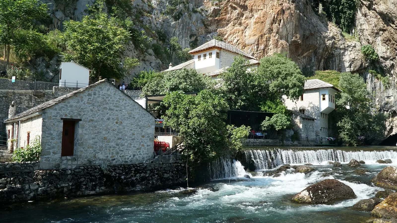 Blagay Tekkesi'nin beyaz badanalı yapıları, arkasındaki sarp kayalık ve önünden akan Buna nehri