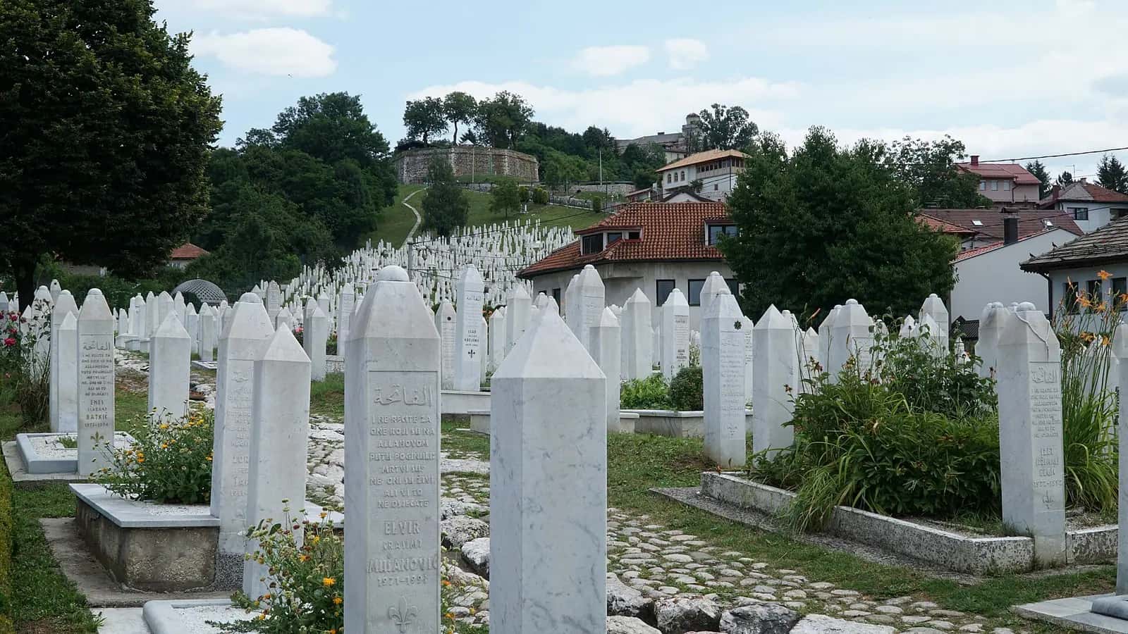 Kovači Şehitliği'nde sıra hâlinde dizilmiş beyaz mezar taşları ve arkadaki evler